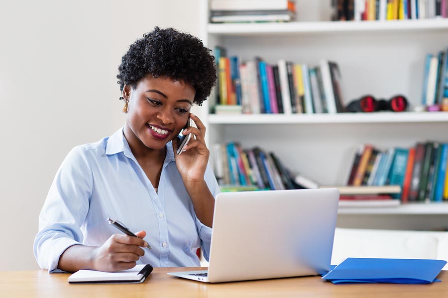 Contact Us - Businesswoman Smiles and Makes a Call While Taking Notes, at Her Desk in a Bright Office